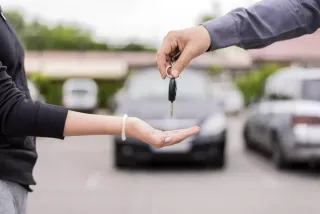 Mano sosteniendo llave de auto y otra recibiéndolas y de fondo un auto 